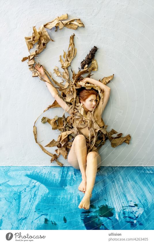 Young sexy woman with red hair artfully covered with dry, withered decorative banana tree leaves sitting on the blue floor. copy space studio floor. copy space
