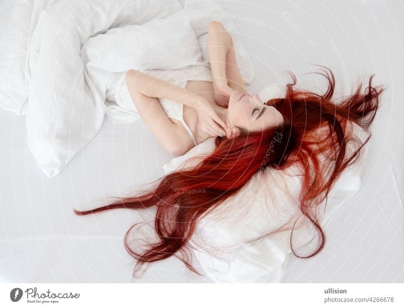 Top view of attractive, contented, young, sexy, woman with dyed bright red hair relaxing in bed, enjoying soft sheets and mattress in bedroom, copy space. Woman