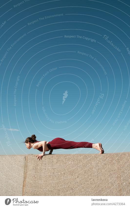 Young brunette woman practicing chaturanga dandasana on top of concrete wall on a beach in front of blue sky yoga yogini plank pose sport athletic female