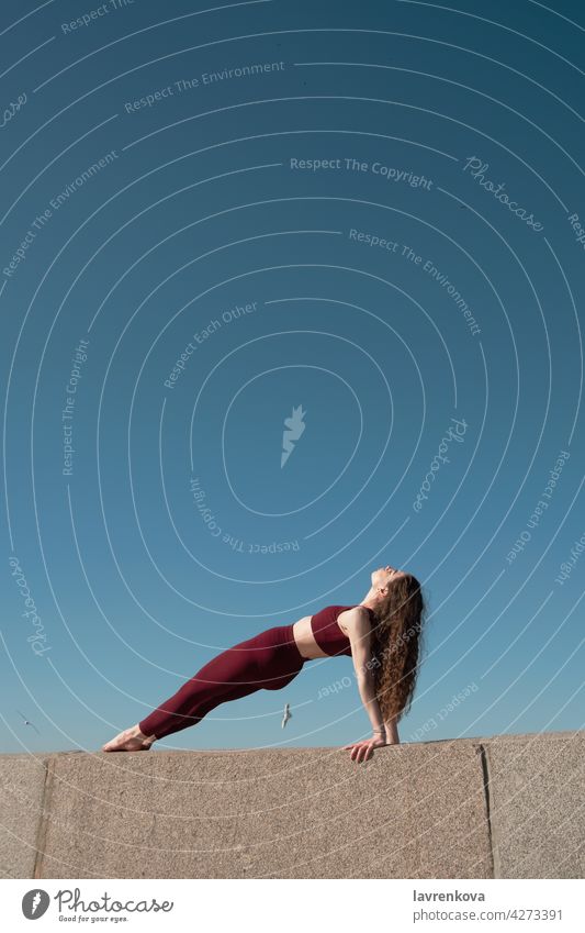 Young brunette woman practicing Purvottanasana pose on top of concrete wall on a beach in front of blue sky Reverse Plank Pose yoga yogini sport athletic female