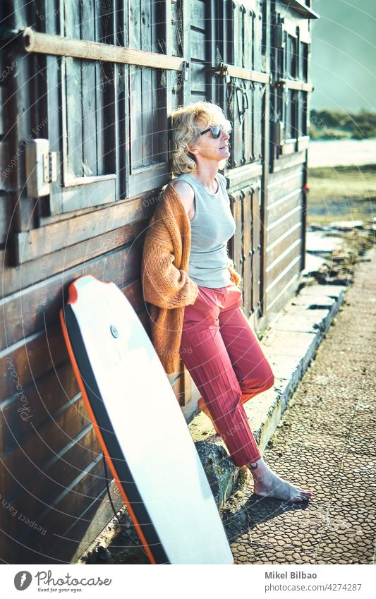 Portrait of a young mature blonde caucasian woman outdoor in a wooden house close to a beach area with a bodyboard and sunglasses.  Lifestyle concept. lifestyle