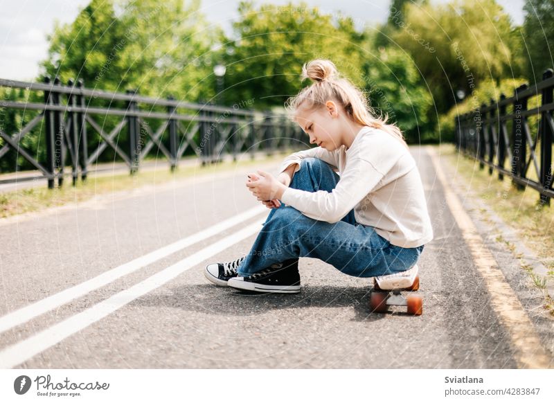 Cute blonde teenage girl sitting on a skateboard in a city park chatting on a smartphone with friends and taking a selfie. skating happy cheerful
