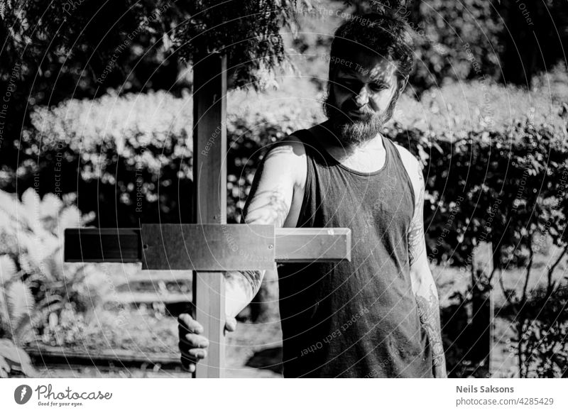 male with tattoos and beard holding wooden christian cross in cemetery. portrait tattoed catholic religion Religion and faith Crucifix Christian cross