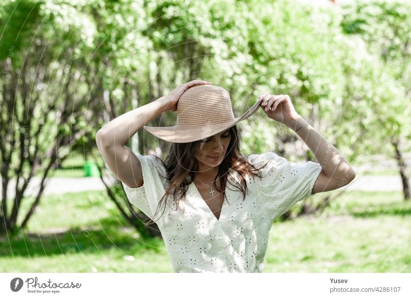 Young beautiful happy woman in fashionable straw hat. A brunette female in a white shirt stand outdoor and holds her hat with hands. Blooming spring apple tree park background. Sunny summer day