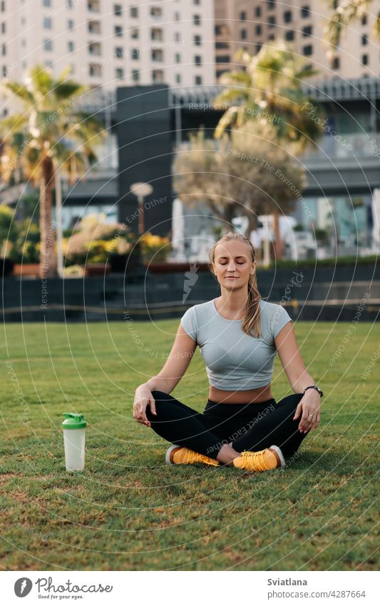 A young woman sits in the lotus position on the lawn in the park next to the house and meditates after a workout. Sports, fitness, yoga. Healthy Lifestyle Concept