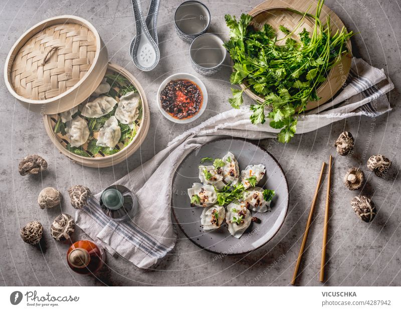 Asian food concept with homemade dumplings on plates and in steamer, fine shiitake, traditional sauces and crockery. White kitchen cloth on grey concrete background. Top view