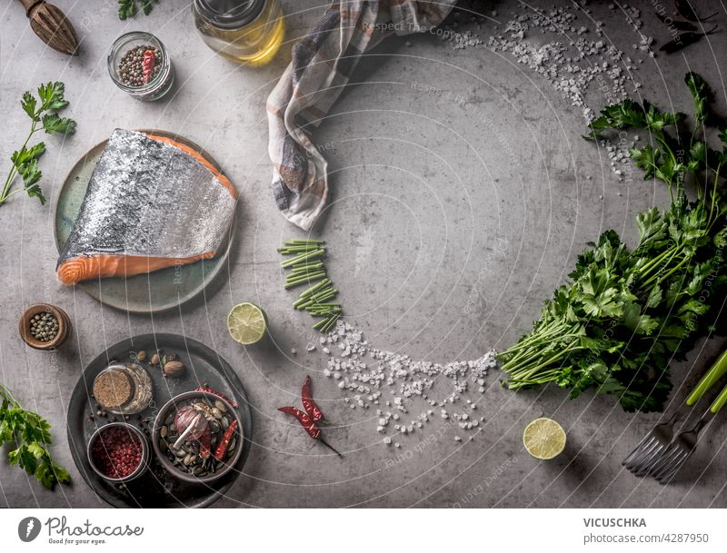 Fresh raw salmon with frame of spices, oil, fresh herbs and lemon on grey concrete background. Cooking preparation with fresh ingredients at home. Equipment: fork, bowls and kitchen cloth. Top view