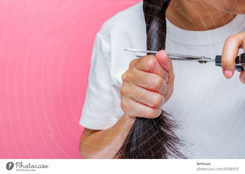 Women cutting damaged and dry hair. Asian woman cutting hair with scissors for donating to cancer patients. Hair donation for breast cancer person. Woman with black long hair on pink background.
