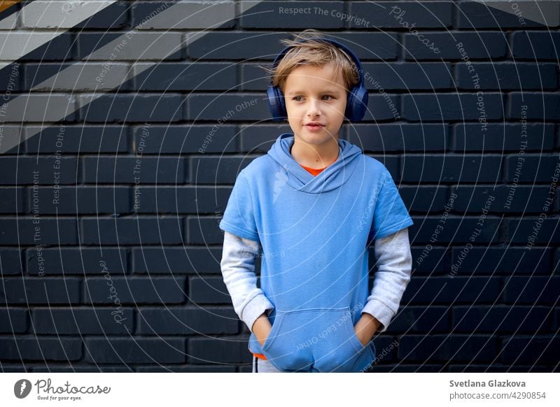 Blonde Caucasian boy in a blue hoodie and headphones stands on a city street against a gray brick wall, listening to music in a good mood lifestyle portrait