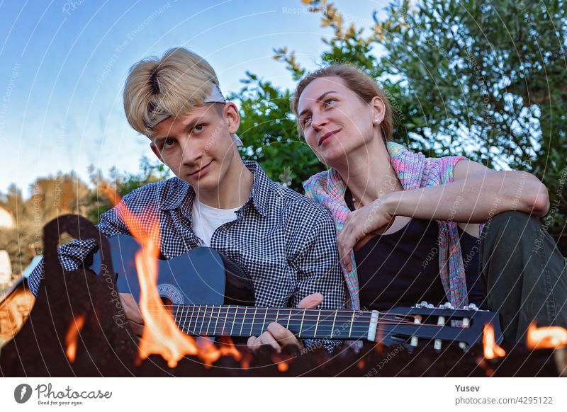 Happy cheerful mom and son sit in the backyard near the fire and play an acoustic guitar. They sing songs and enjoy a summer evening. Summer lifestyle photography. Blue sky background.