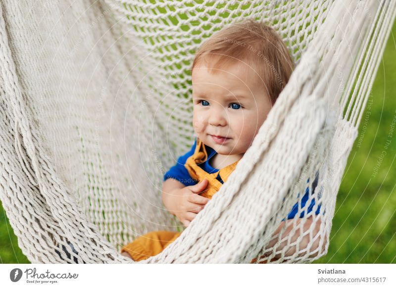 A little boy sits in a hammock on a summer day in the garden baby toddler child sun green nature summertime sunbathing relax outside park happy sitting cute