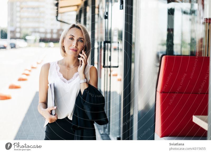 A beautiful businesswoman, talking on the phone in the city, holding a folder with documents street portrait standing girl lifestyle executive young success job