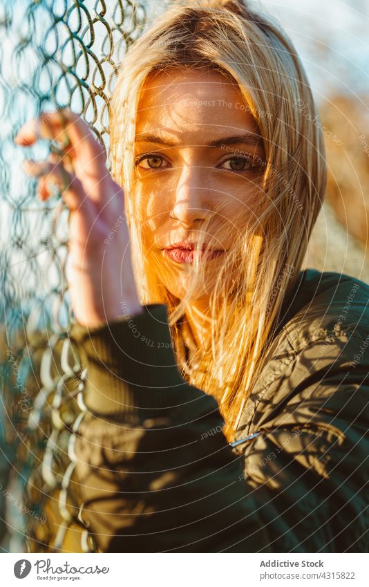 Calm woman leaning on net fence in sunlight portrait chain link calm blond charming modern beauty young bright carefree alone jacket long hair style natural