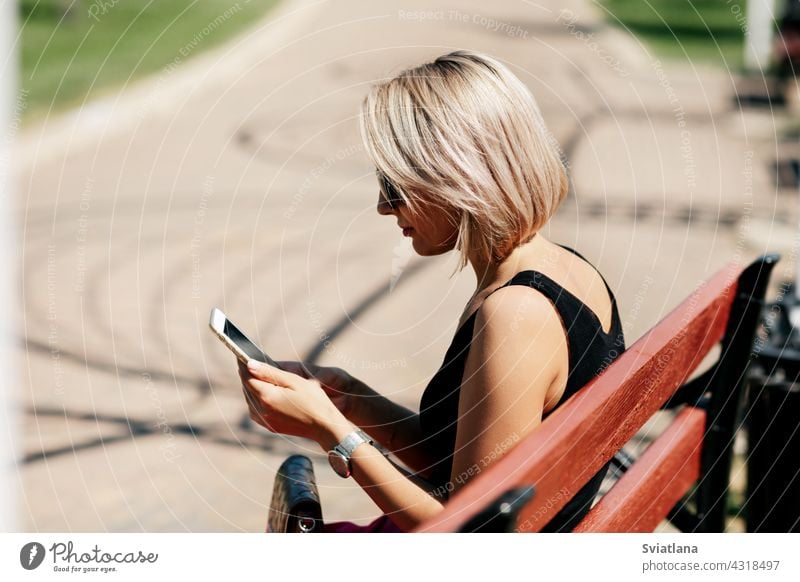 A young woman takes a selfie sitting on a park bench, adjusts her makeup and hairstyle, looking at her reflection on the screen taking phone portrait happy girl