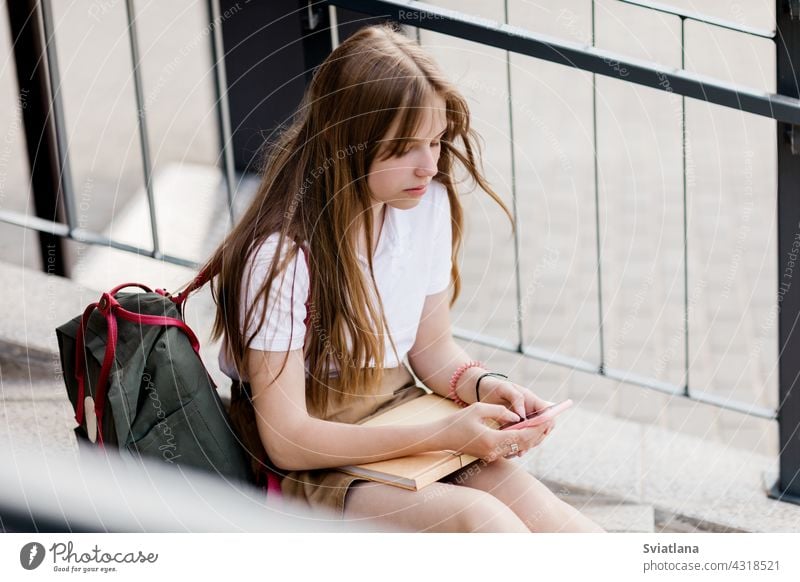 Cute girl sitting on the stairs outside the campus after college class and chatting with her friends teenager phone using backpack school book messaging young