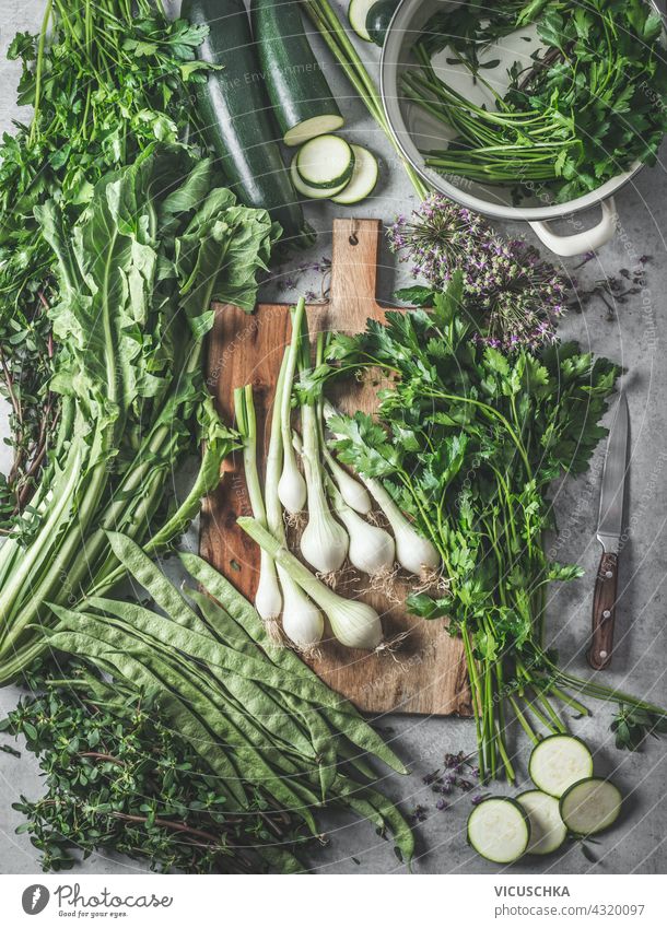 Variety of green vegetables and herbs on a rustic kitchen table with a wooden cutting board and pans for healthy cooking at home. Top view. variety top view