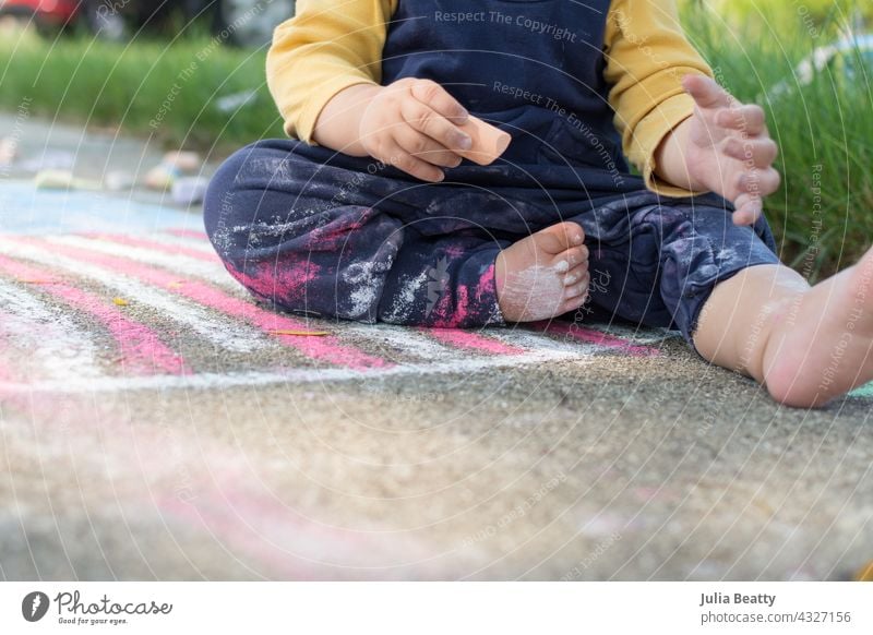 13 month old baby holding a piece of sidewalk chalk, toddler sits on a drawing of the American Flag and has chalk dust on pants and body 4th of july