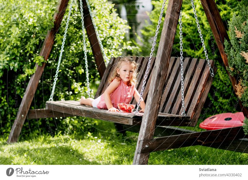 A little girl is lying on a garden swing and eating fresh strawberries baby strawberry lie down toddler summer cute fun fruit spring sunny colorful day green