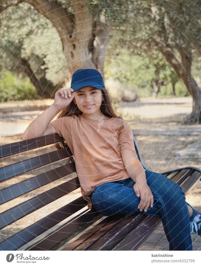 Smiling girl kid wearing t-shirt; jeans and baseball cap smile casual mock up bench park baseball hat child copy space Person Portrait preteen alone round neck