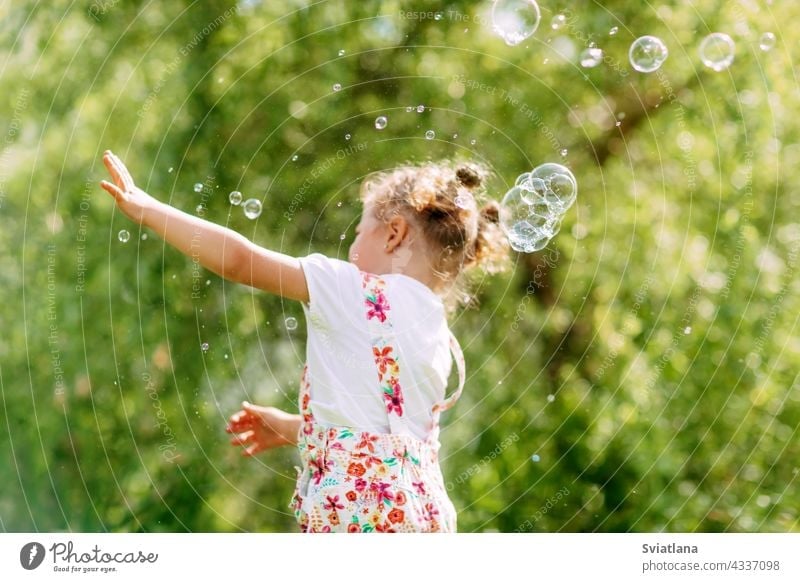 A cheerful girl catches soap bubbles with her hands. Happy childhood, summer time. Side view little joy baby green fun portrait blow beautiful kid park play