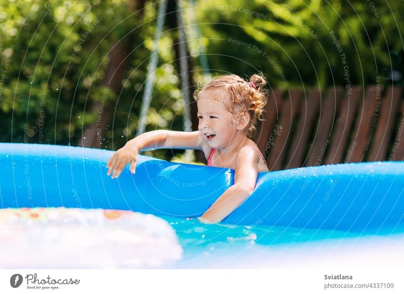 A charming baby touches the water in an inflatable pool in the garden and smiles girl playing fun childhood summer kid swimming joy swimsuit family funny little