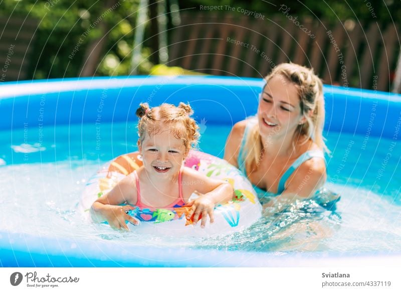 A young mother teaches her little daughter to swim in the pool in the garden with the help of an inflatable circle mom swimsuit water childhood cheerful weekend