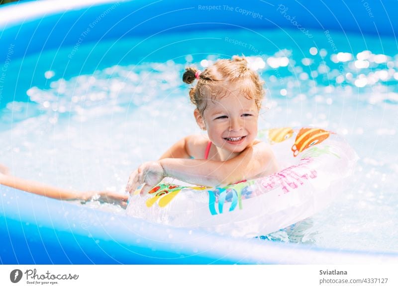 A laughing baby in a bright swimsuit learns to swim in the pool in the garden with the help of an inflatable circle. Summer time, recreation, rear view girl