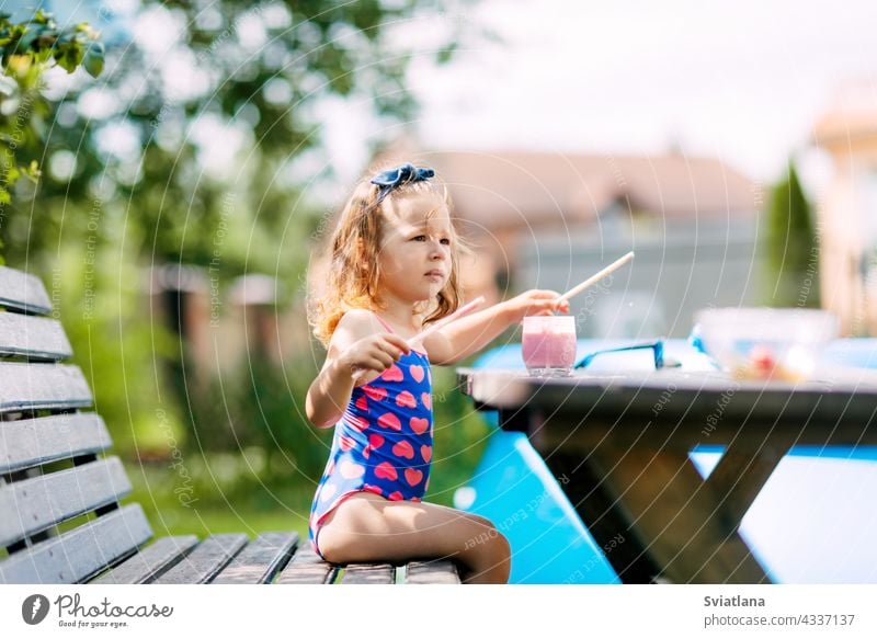 A cute baby is drinking a berry smoothie through a straw, sitting on a bench in the garden. Outdoor recreation, summer holidays for children healthy vitamin