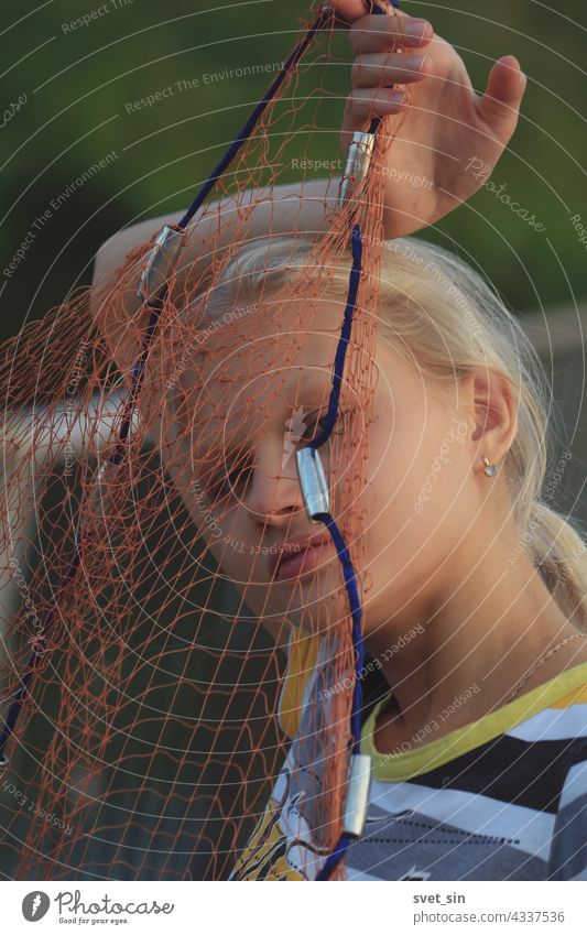 Portrait of a blond teenage girl through a fishing net. A blond girl holding a fishing tackle in front of her face outdoors. blonde fishnet teenager portrait