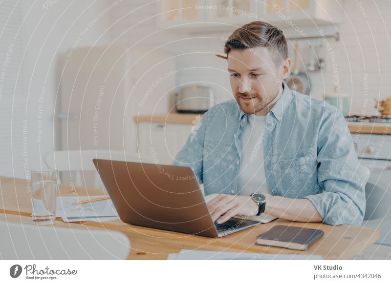 Focused young man freelancer using laptop while sitting at kitchen table working remotely home data handsome smile indoors male business computer casual typing