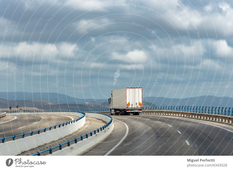 Truck with metal semi-trailer, widely used in textile and automotive industry, driving over bridge with cloudy sky. Trailer Street Transport Highway Cargo