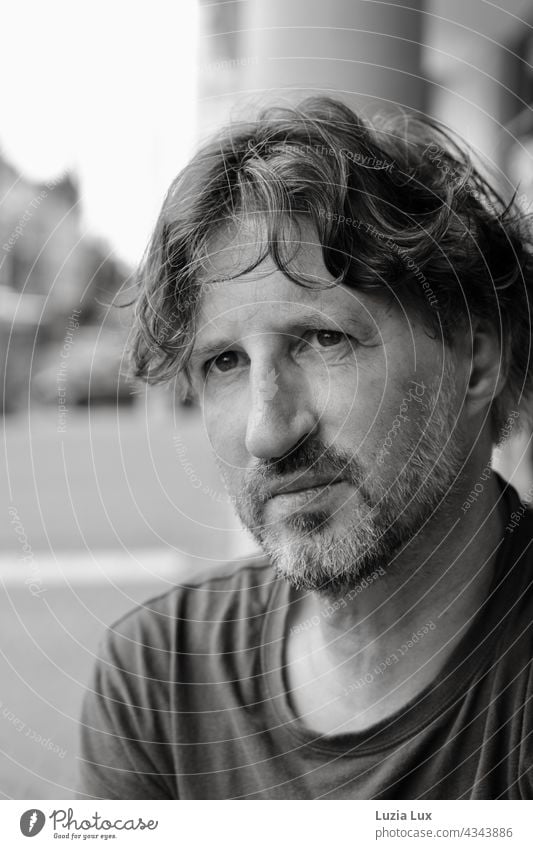 A curly-haired, middle-aged man sits in front of a sidewalk café, looks into the camera Grinning Disheveled Curl Designer stubble Facial hair Adults kind