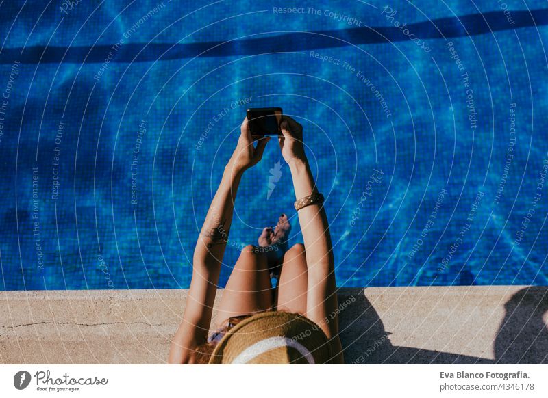 top view of young caucasian woman sitting by pool side using mobile phone to take a selfie. Summer time, vacation and lifestyle technology swimming pool