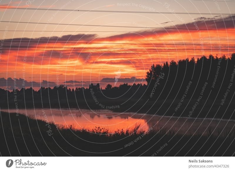 sunset over the river. Red orange clouds. Black forest silhouette over the river. Electric power line wires in he air. Reflection of clouds and reeds in river makes it orange