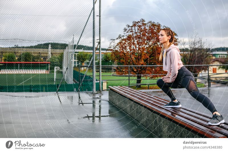 Young sportswoman stretching legs outdoors on a bench self-confident athlete copy space body warmup training crouched exercising rainy morning sporty