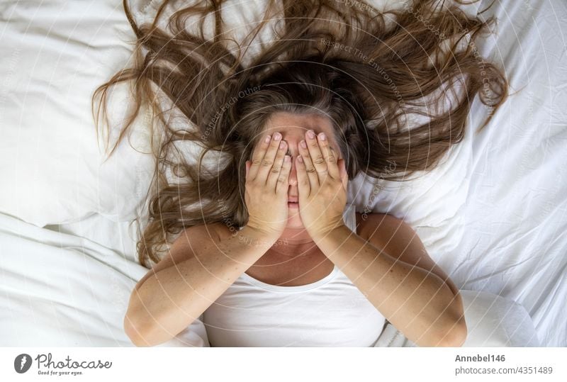 Young woman lying in bed suffering, tired woman covering face with hands, can't sleep feels exhaustand on white sheets in bedroom head ache bedroom closeup