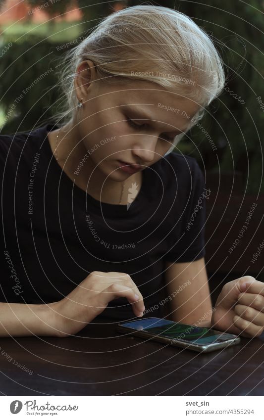 A blond teenage girl in a black T-shirt is sitting outdoors at a wooden table and flips through (reads) posts in social networks on her smartphone. blonde