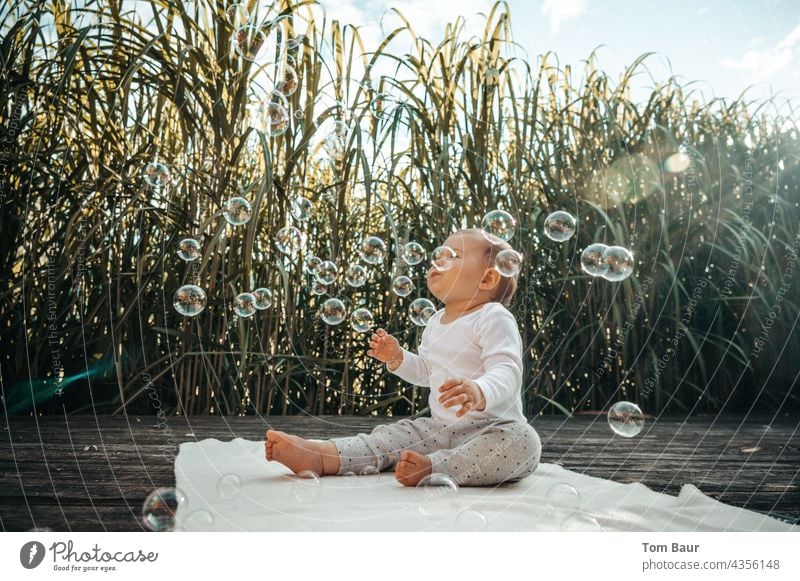 Baby sits on the floor in front of reeds and plays with soap bubbles Soap bubble Playing Joy Infancy Happy Child Happiness Blow Sky Common Reed Exterior shot