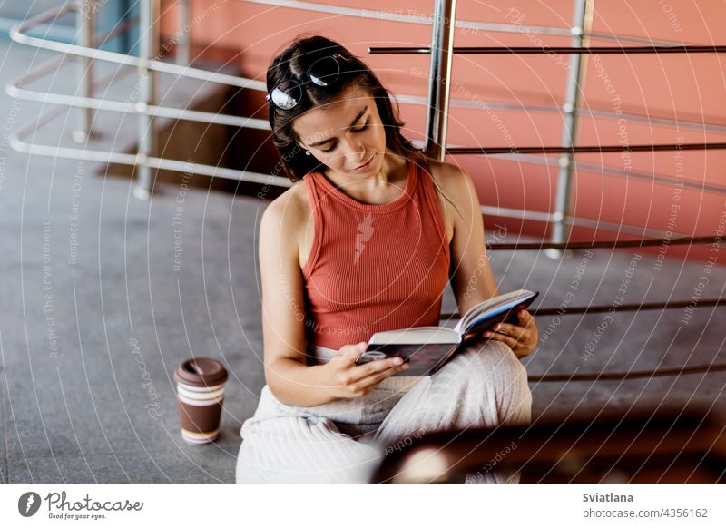 A student sits on the stairs on campus and reads a book, next to her is a glass of coffee girl sitting reading learning college school beautiful lifestyle day