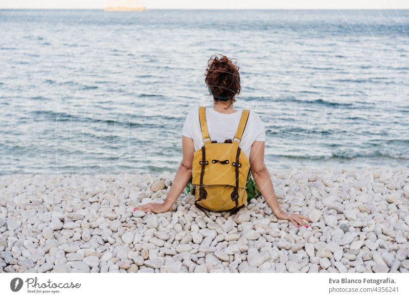 back view of relaxed caucasian woman with yellow backpack sitting by the beach during sunset. summer time. daydreaming. Outdoors lifestyle ocean sea holidays