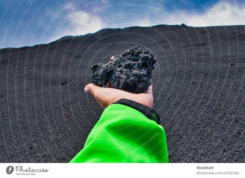 Etna - woman's hand holding a large volcanic ash lump in her outstretched hand towards the volcanic crater Women`s hand green jacket blue sky white clouds