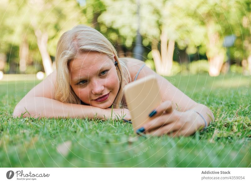Glad woman taking selfie and listening to music on green lawn in park earphones summer entertain song smile enjoy female lying grass happy relax smartphone