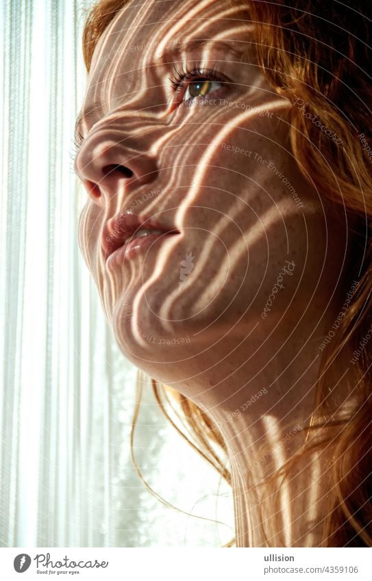 Portrait of a beautiful young attractive sexy redhead woman, smiling seductively, shadow striped by the threads of a string curtain in the early morning sun