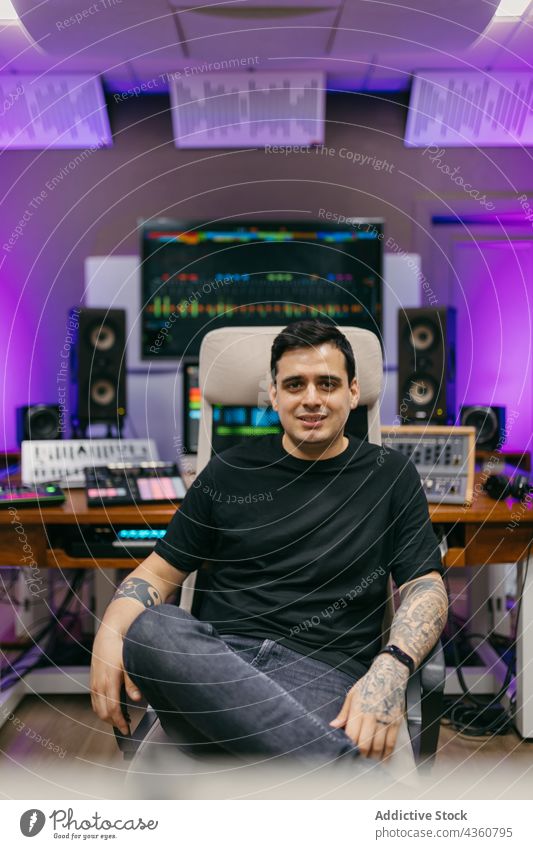 Sound man with tattoos in music studio with neon light engineer sound confident legs crossed professional portrait self assured smile equipment modern style
