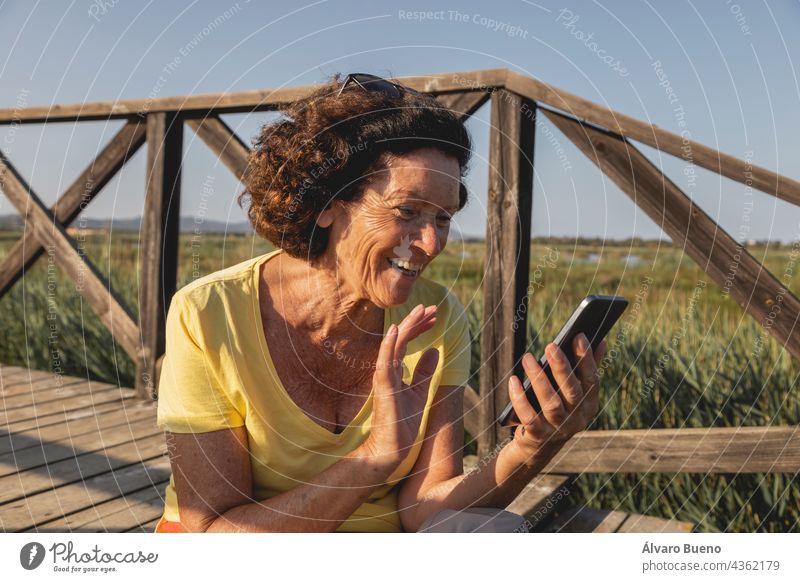 A cheerful and vital woman, in her 70s, greets while chatting with someone on a video call from a natural area of the Mediterranean, Spain waving saying hello