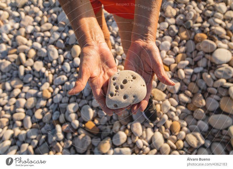 A woman, in her 70s, holds a stone full of holes, on a natural beach, next to the Mediterranean Sea, Spain texture object pebble beach hands close-up senior
