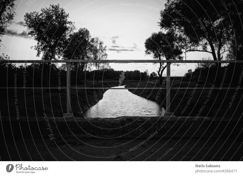 black and white landscape of river and some trees talen from old bridge. Clear sky with some clouds. Bright water. Bridge iron railing on foreground. Rectangles.