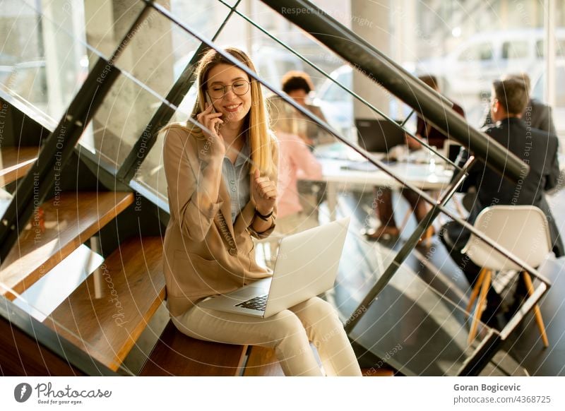 Young woman sitting on the stairs with laptop and using mobile phone in modern office in front of her team adult attractive beautiful business businesswomen