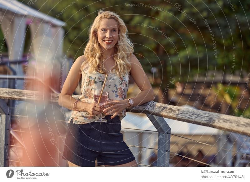 Pretty relaxed blond woman enjoying a tropical cocktail on summer vacation standing leaning on wooden railings looking at the camera with a friendly smile and copyspace