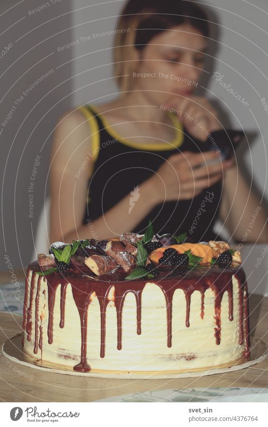 A birthday cake decorated with chocolate, berries, fruits, mint leaves, and a girl reading messages on a mobile phone in the background. Indoors. berry leaf